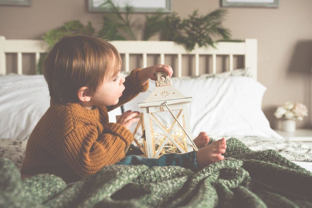 Toddler in a knitted jumper sitting in bed looking at a lantern with a warm glow