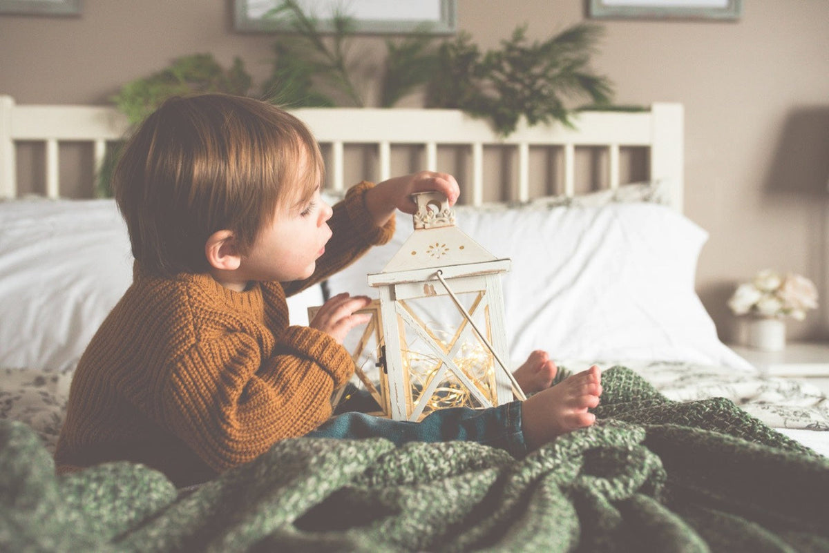 Toddler in a knitted jumper sitting in bed looking at a lantern with a warm glow
