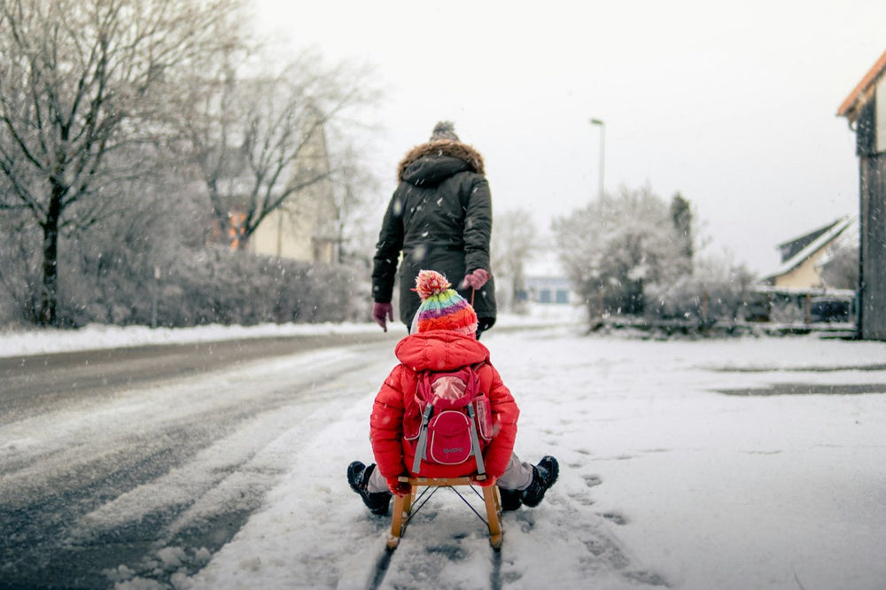 A child in a red winter jacket and striped hat sits on a sled pulled by an adult along a snowy street